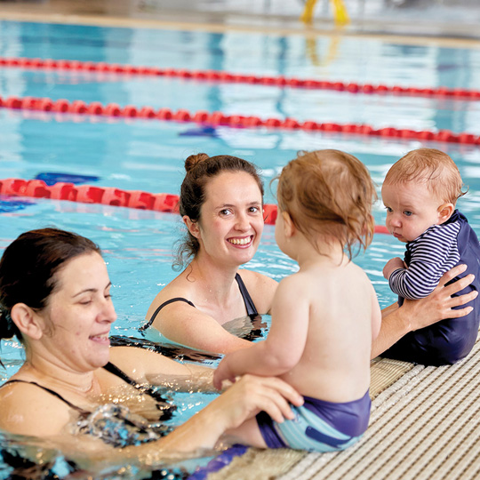 Parents in water holding babies siting on the edge of an indoor pool